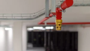 Close-up of a brass clean agent fire suppression nozzle connected to red piping on a server room ceiling, with blurred data center server racks in the background.